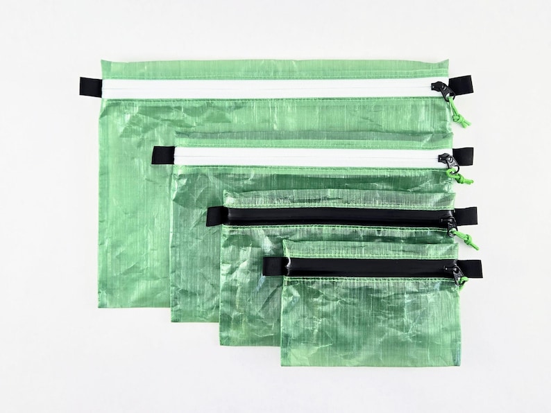 Op de afbeelding: Een set van vier rechthoekige etuis in verschillende maten, gemaakt van een gekreukt, doorschijnend lichtgroen materiaal. Elk etui is voorzien van een horizontale ritssluiting met een zwarte trekker en een klein groen koordje.