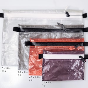 May include: Four zippered pouches in varying sizes and colors. The largest is clear, the next is gray, then orange, and the smallest is purple. Each pouch has a black zipper and pull tab, with measurements in inches and grams.