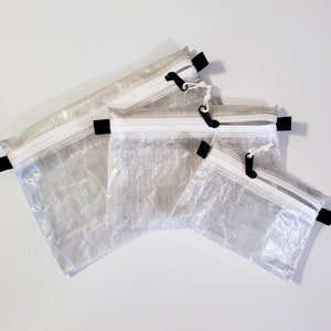 Op de afbeelding: Een set van drie rechthoekige, semi-transparante tasjes gemaakt van gekreukt, wit materiaal. Elk tasje is voorzien van een witte ritssluiting met een zwarte trekker en kleine zwarte stoffen lussen aan de zijkanten om op te hangen.