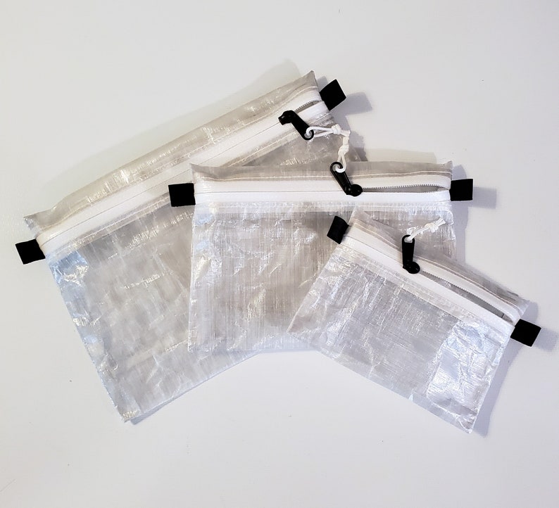Op de afbeelding: Een set van drie rechthoekige, semi-transparante tasjes gemaakt van gekreukt, wit materiaal. Elk tasje is voorzien van een witte ritssluiting met een zwarte trekker en kleine zwarte stoffen lussen aan de zijkanten om op te hangen.