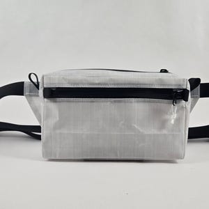 May include: A rectangular waist bag made of a light grey, semi-translucent material with a grid-like texture. It features a horizontal black zippered pocket on the front, a black top zipper, and an adjustable black strap with a plastic buckle.