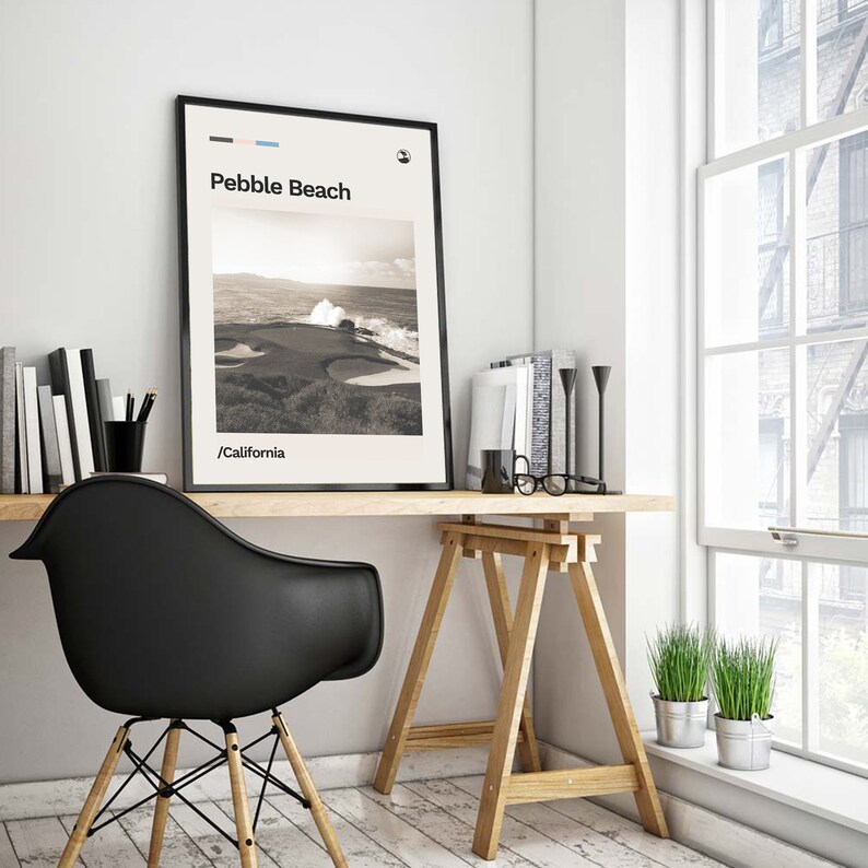 May include: A black and white photo of a golf course with the text "Pebble Beach /California" on a white background. The photo is framed and hanging on a white wall above a wooden desk with a black chair and a wooden easel.