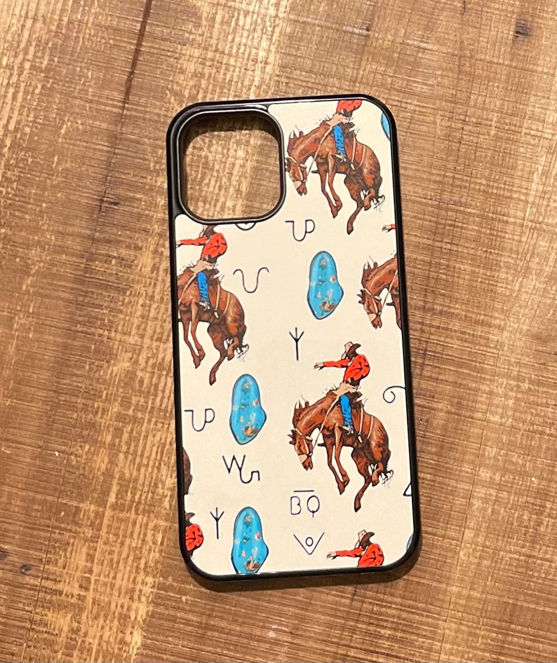 May include: A white phone case with a black border and a pattern of cowboys riding horses and turquoise stones. The case has a cutout for the camera lens and a black button on the side.