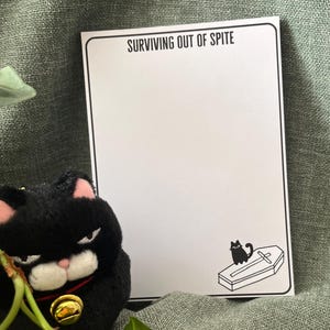 May include: A white notepad with the text "SURVIVING OUT OF SPITE" and a small illustration of a cat on a coffin. A black cat plush with a gold bell sits next to the notepad. Green leaves and a green fabric background.