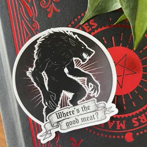 Pode incluir: Um autocolante a preto e branco com um lobisomem e o texto "Where's the good meat?". O autocolante é redondo com uma borda branca e um fundo gradiente vermelho. O autocolante é colocado num livro escuro com detalhes vermelhos.