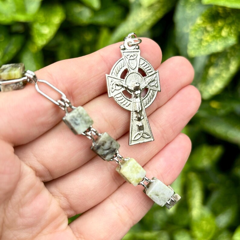 Pu&ograve; includere: Un ciondolo a forma di croce celtica in argento con un crocifisso e un intricato disegno di nodi. Il ciondolo &egrave; attaccato a una collana del rosario con perline di pietra verde e bianca.