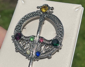 May include: Silver Celtic knot brooch with a sword design and colorful gemstones. The brooch features a circle with a sword in the center and six gemstones in various colors, including green, red, blue, yellow, and purple.