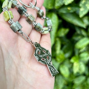 Pu&ograve; includere: Una collana con catena d'argento con un ciondolo a croce celtica e perline di pietra verde e bianca.