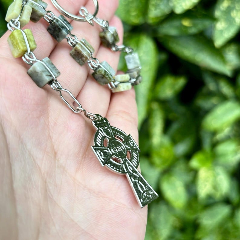 Pu&ograve; includere: Una collana con catena d'argento con un ciondolo a croce celtica e perline di pietra verde e bianca.