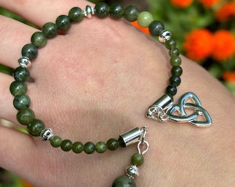 May include: A green jade stone bracelet with a silver Celtic knot charm and a silver bead toggle clasp. The bracelet is on a person's wrist.