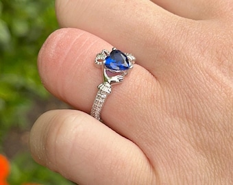 May include: A silver ring with a blue heart-shaped stone. The ring is a Claddagh ring, a traditional Irish design featuring two hands clasping a heart.