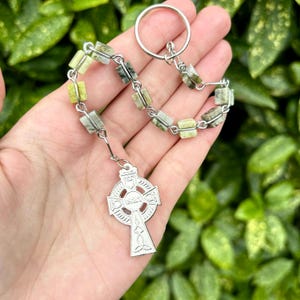 Pu&ograve; includere: Un ciondolo a forma di croce celtica argentata con una catena di rosario in pietra verde e bianca. La catena ha un anello d'argento nella parte superiore.