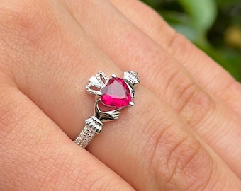 May include: A silver ring with a heart-shaped red gemstone. The ring is a Claddagh ring, a traditional Irish design featuring two hands clasping a heart.