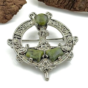May include: A silver Celtic knot brooch with green stone accents. The brooch features a circular design with intricate details and a central stone.