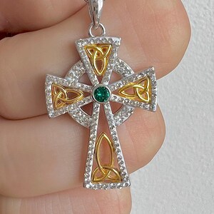 Peut inclure: Un pendentif en forme de croix celtique argentée et dorée avec une émeraude verte au centre. La croix est décorée d'un motif de nœud et de strass transparents.