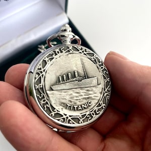 Puede incluir: Reloj de bolsillo plateado con una imagen en relieve del Titanic. El reloj tiene un borde adornado y un vástago de cuerda. La palabra "TITANIC" está impresa en la esfera. El reloj se presenta en una caja forrada de terciopelo verde.