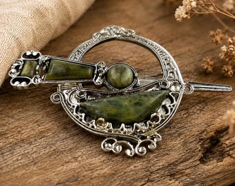 Può includere: Una spilla in metallo color argento caratterizzata da una cornice circolare con intricati motivi a volute e tre pietre lisce incastonate di colore verde oliva. Il design include una spilla orizzontale che attraversa il centro della cornice.