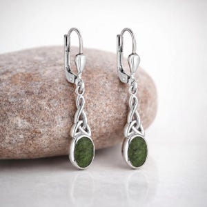 May include: A pair of silver drop earrings featuring a polished oval green stone set at the base. The earrings have a leverback closure and a decorative metal knot design connecting the hook to the stone setting, creating a balanced appearance.