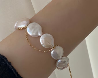 Puede incluir: Un brazalete de cadena dorada con grandes perlas barrocas blancas. Las perlas tienen una forma irregular y un acabado liso y brillante.