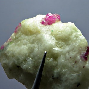 Könnte beinhalten: Nahaufnahme eines weißen, grob strukturierten Gesteins mit einem Cluster aus rosafarbenen Rubinkristallen auf der Oberseite. Das Gestein wird von einem hellen Licht beleuchtet, das den Glanz der Kristalle hervorhebt.