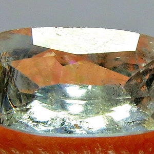 May include: A faceted, rectangular gemstone with rounded corners. The gem is primarily a pale blue-green color with internal reflections of orange and pink. The stone is held by a finger, suggesting it is a loose gemstone.