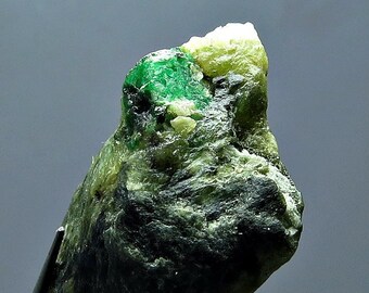 Könnte beinhalten: Ein roher Smaragdedelstein mit leuchtend grüner Farbe, akzentuiert durch hellere grüne und cremefarbene Bereiche. Der unbearbeitete Rohstoff wird von einer Pinzette gehalten, die seine natürliche, unpolierte Textur und Kristallstruktur zeigt.