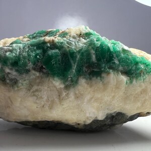 以下が含まれることがあります： 鮮やかな緑色のエメラルドの結晶が、質感のあるオフホワイトの大きな石英マトリックスに埋め込まれた、原石の標本。石の底部は濃い灰色で、明るい上部と対照的なコントラストを生み出しています。