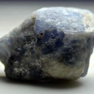 May include: A rough, blue-grey gemstone with a natural, irregular shape. The stone has a cloudy, opaque appearance with a slightly textured surface.