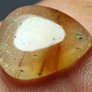 Peut inclure: Une pierre précieuse translucide en forme de goutte d'eau, avec une zone blanche centrale et une bordure ambrée. La pierre présente des inclusions sombres et de petits points verts, avec une surface polie.