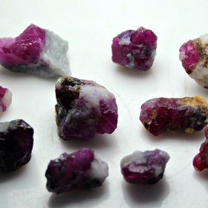 May include: A collection of rough ruby gemstones, showcasing a vibrant red hue with white and gray matrix. The raw stones vary in size and shape, displaying natural textures and crystalline structures. The gemstones are scattered on a white surface.
