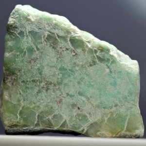 Puede incluir: Un espécimen de piedra natural en bruto, con una superficie moteada y texturizada. La piedra muestra una mezcla de tonos verdes y verde azulado pálido, con sutiles variaciones de tono. La forma general es irregular, mostrando la naturaleza cruda y sin pulir del mineral.