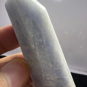 May include: A pale blue, elongated crystal specimen with a translucent, semi-opaque appearance. The mineral features a naturally faceted, pointed termination at one end and a textured, uneven surface with subtle white and light blue color variations.