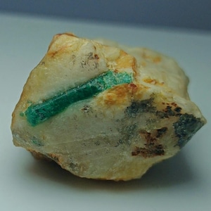 Puede incluir: Un cristal de esmeralda natural en bruto incrustado en una matriz de roca beige y marrón claro. La esmeralda es de un color verde intenso y vibrante, que contrasta con los tonos más claros de la piedra circundante. La roca tiene zonas grises y marrones.