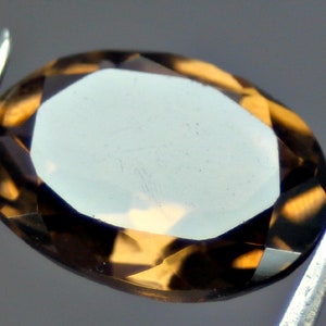 Könnte beinhalten: Ein oval geschliffener Edelstein mit facettierter Oberfläche, gehalten von einer silbernen Pinzette. Der Edelstein hat einen hellbraunen Farbton mit dunkleren braunen Facetten, die das Licht reflektieren. Geeignet für die Schmuckherstellung.