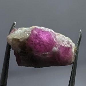 Może przedstawiać: Surowy, nieoszlifowany kamień szlachetny rubinu, trzymany przez metalową pęsetę. Rubin ma żywy różowo-czerwony odcień, z szorstką, teksturowaną powierzchnią. Kamień jest częściowo osadzony w jaśniejszej matrycy.