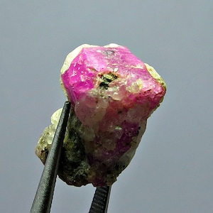 May include: A vibrant, raw ruby gemstone with a striking pink hue, held by tweezers. The ruby is partially embedded in a matrix of lighter-colored rock with hints of green. The gemstone's surface has a rough, natural texture.