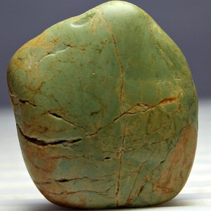 May include: A smooth, rounded stone with a mottled green and brown surface. The stone has a polished appearance with visible cracks and variations in color, suggesting natural formations. The overall shape is irregular and organic.