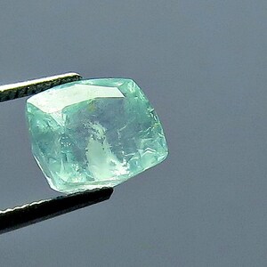 May include: A faceted, light blue-green gemstone held by tweezers. The gem has a slightly rounded, square shape with multiple facets that catch the light. The background is a solid, muted gray.