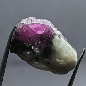 Pode incluir: Uma gema bruta, não cortada, segurada por uma pinça. A gema apresenta um vibrante núcleo cristalino rosa, que se desvanece para um exterior mais claro e opaco. A textura áspera e a forma natural da pedra são claramente visíveis.