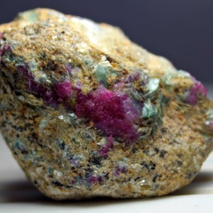 Op de afbeelding: Een ruwe, bruine steen met ingebedde roze en groene kristallen. De kristallen zijn verspreid over de hele steen en creëren een kleurrijk en interessant patroon.