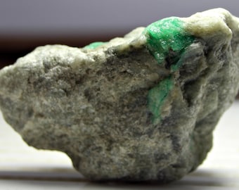 May include: A rough, gray rock with a bright green emerald crystal embedded in it.