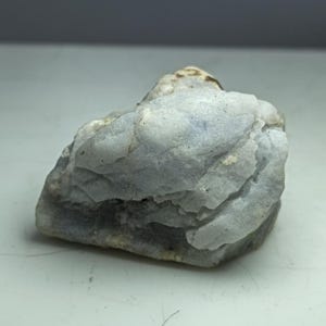May include: A rough, irregularly shaped rock with a light blue and gray color palette. The stone has a textured surface with visible ridges and variations in tone, suggesting a natural mineral formation. The overall appearance is of a solid, unpolished specimen.