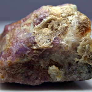 Può includere: Una roccia grezza di forma irregolare con un motivo di colore viola e bianco. La roccia ha una superficie ruvida e testurizzata e sembra essere un campione minerale naturale.