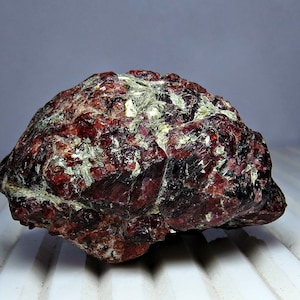 May include: A rough, uncut gemstone with a deep red hue, interspersed with lighter, crystalline patches. The rock's surface is textured and irregular, showcasing natural mineral formations. The overall shape is rounded and compact, with a dark, almost black, base.