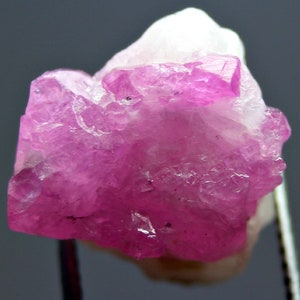 May include: A close-up of a rough, uncut gemstone. The stone is a vibrant pink colour, with areas of white at the top. The crystalline structure is visible, with sharp edges and a textured surface. The gemstone is likely a mineral specimen.