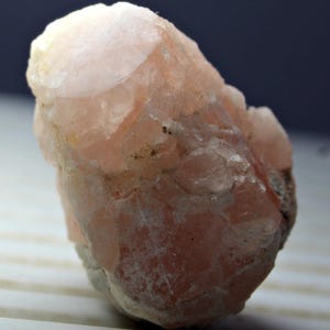 May include: A raw, uncut mineral specimen with a pale pink hue. The crystalline structure is visible, with a rough, textured surface. The stone has a soft, almost translucent appearance, with areas of lighter and darker pink tones. The overall shape is irregular and natural.