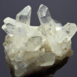 Könnte beinhalten: Ein Cluster aus klaren Quarzkristallen auf einer rauen, grauen Matrix. Die Kristalle sind scharf und spitz, mit einem glasartigen Glanz.