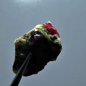 Könnte beinhalten: Ein roher Edelstein, oben leuchtend rot, übergehend in Grün und Weiß. Der Stein wird von einer Pinzette gehalten, wodurch seine natürliche, ungeschliffene Textur hervorgehoben wird. Der Hintergrund ist ein sanftes Grau.