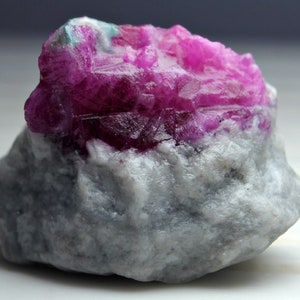 Puede incluir: Una gema en bruto, sin cortar, con una vibrante formación de cristales de color fucsia sobre una matriz gris y blanca. La estructura cristalina muestra facetas afiladas y angulares, contrastando con la base rugosa y texturizada. La muestra mineral es una roca natural sin pulir.