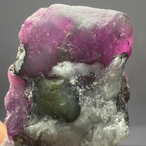Könnte beinhalten: Nahaufnahme eines rohen, ungeschliffenen Edelsteins. Der Stein hat oben eine leuchtende magenta Farbe, die in helleres Rosa und Weiß übergeht. Grüne und graue Einschlüsse sind sichtbar, was ein natürliches Aussehen erzeugt.